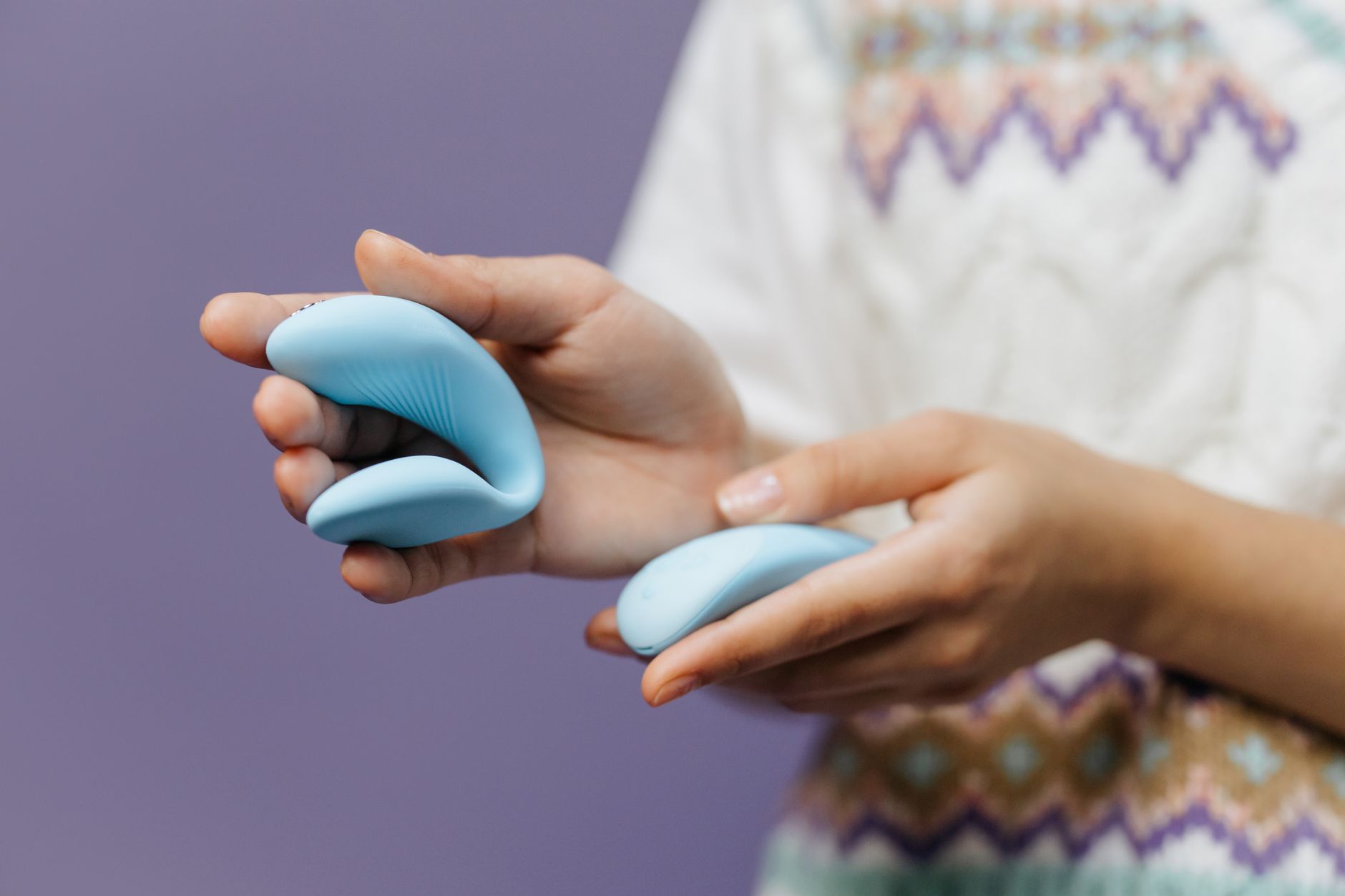 Hands holding a sleek blue clitoral vibrator against a purple background, representing intimate pleasure tools.